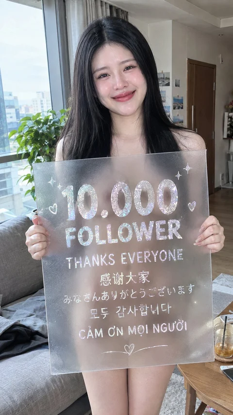 A woman holds a translucent sign celebrating 10,000 followers inside a bright room.